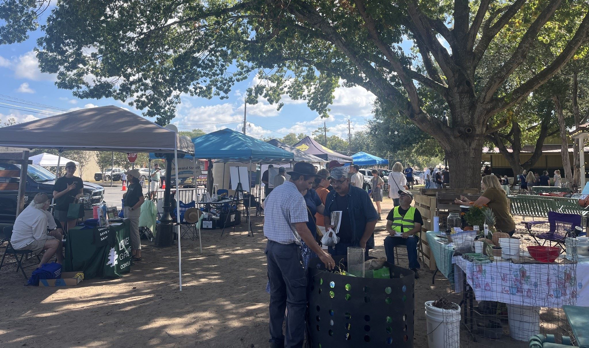 Outdoor community event with people visiting vendor booths under tents and trees.