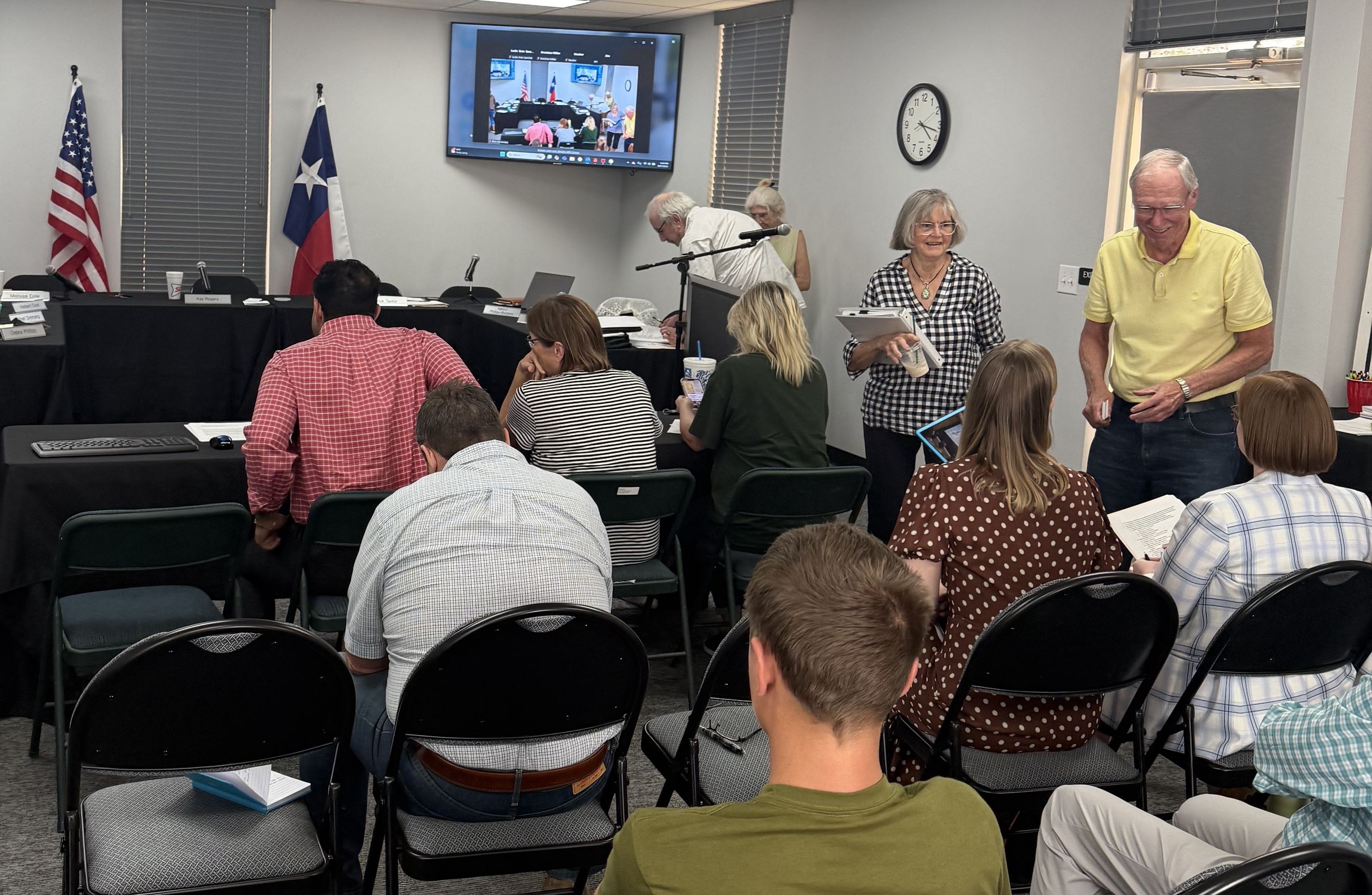 People gathered in a meeting room with chairs, papers, and a screen showing a video call.