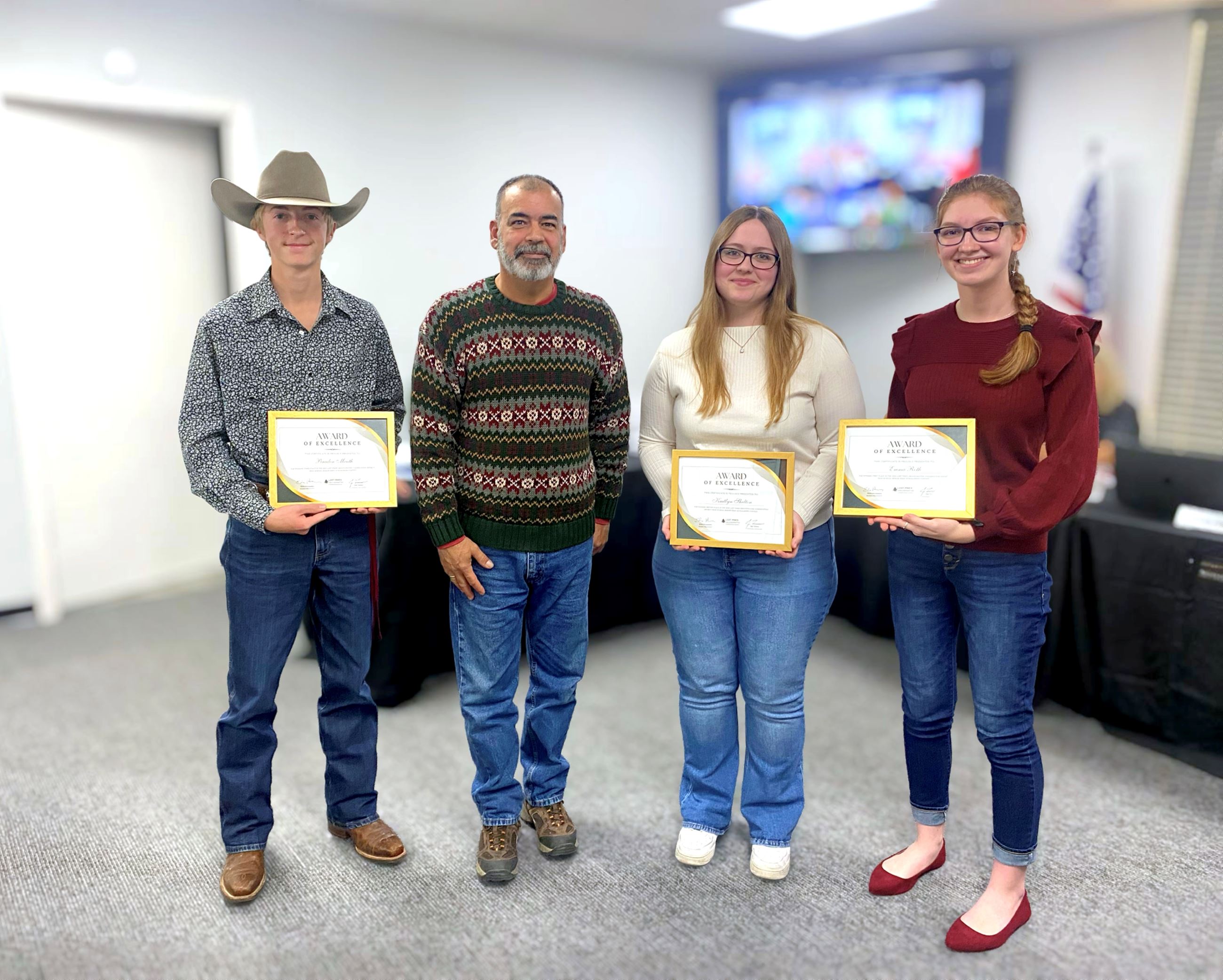 Four people stand together indoors; three hold “Award of Excellence” certificates while the man in t